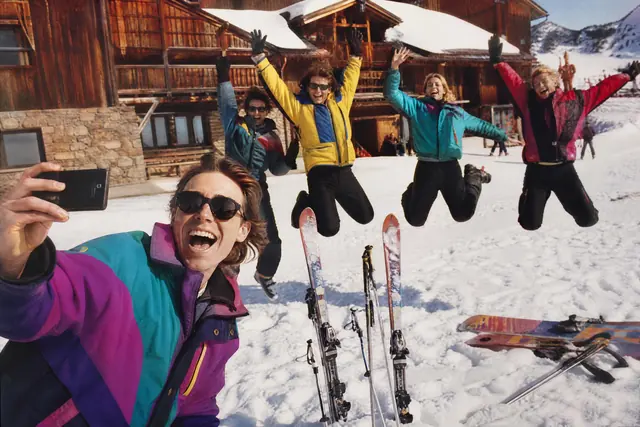 Eine Gruppe von Freunden in Skikleidung springt vor einer Hütte in die Luft, während einer ein Selfie macht